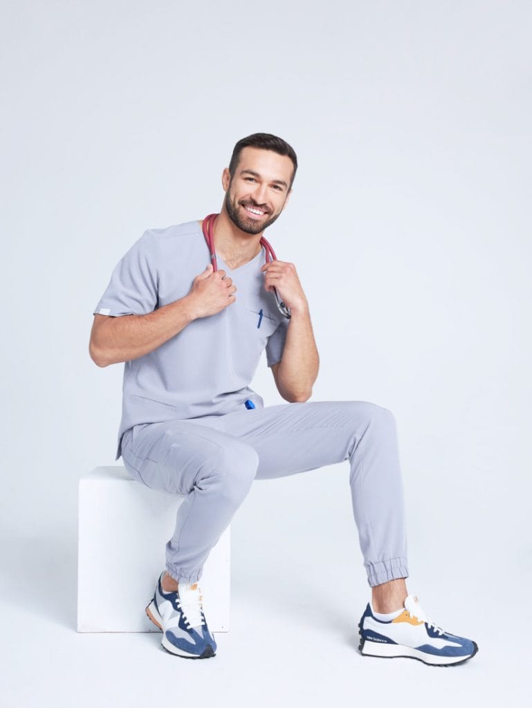 Uśmiechnięty mężczyzna w szarym stroju medycznym z czerwonym stetoskopem siedzi na białym sześcianie.