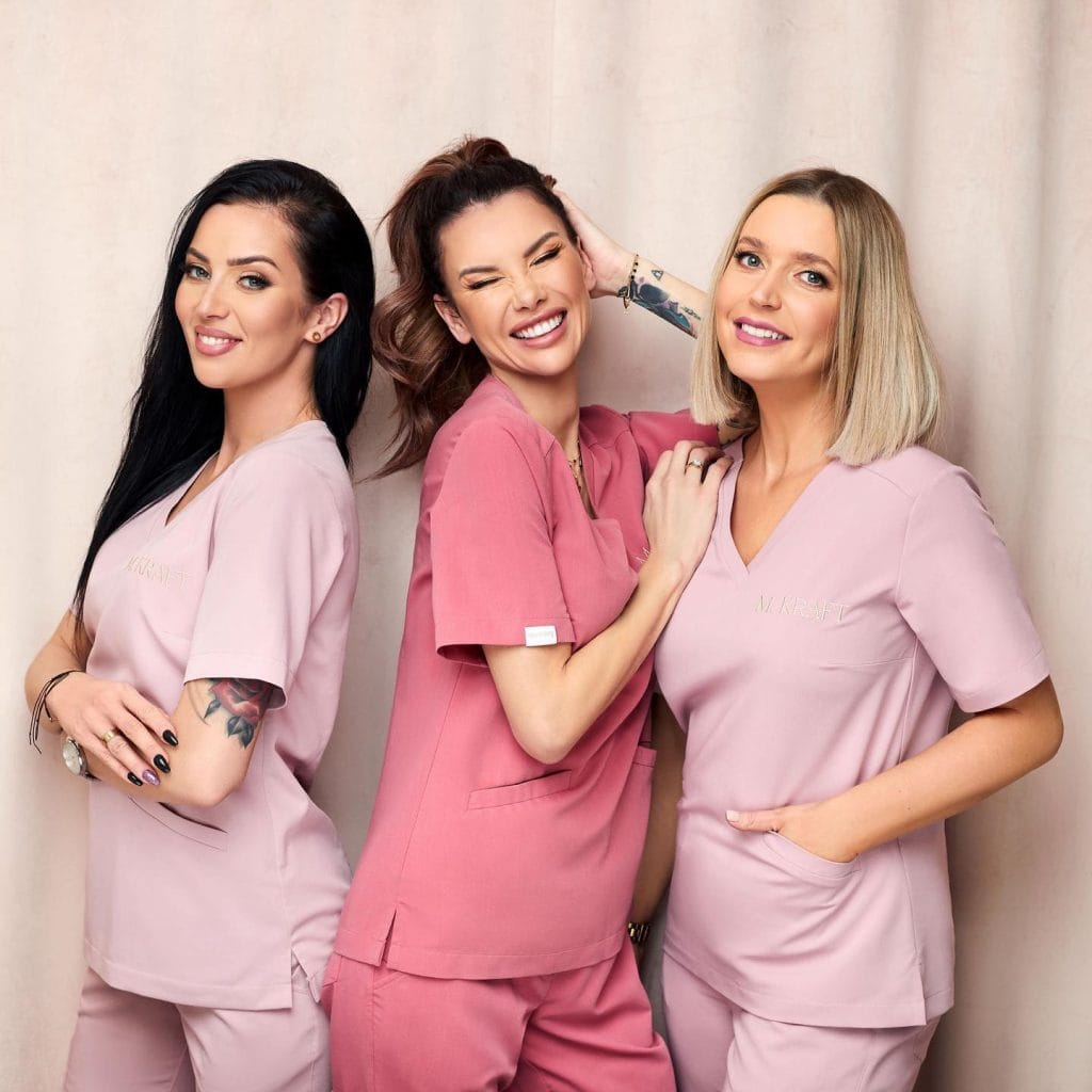 Trzy uśmiechnięte kobiety w kolorowej odzieży medycznej na jasnym tle.