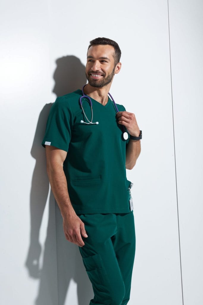 Mężczyzna w zielonym ubraniu medycznym ze stetoskopem uśmiecha się, stojąc przy ścianie.