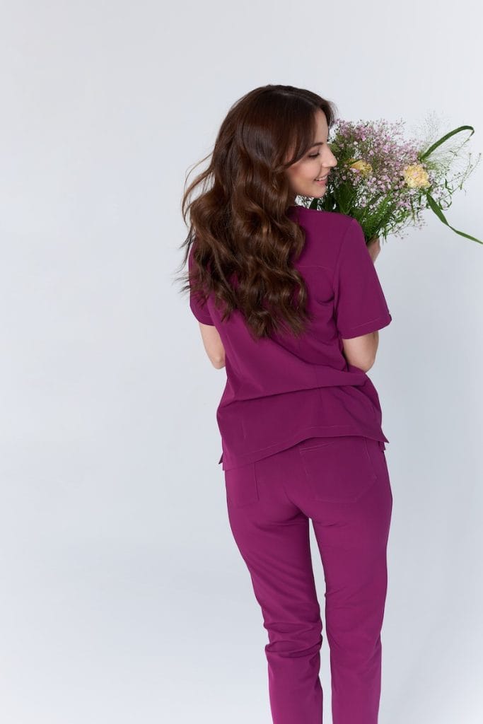 Kobieta w medycznym kompletcie scrubs Basic PLUM z tyłu trzymająca bukiet kwiatów.