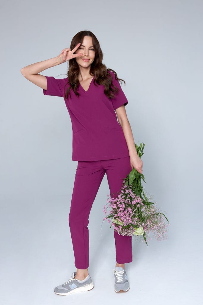 Kobieta w fioletowym medycznym komplecie scrubs trzyma bukiet kwiatów i robi znak pokoju.