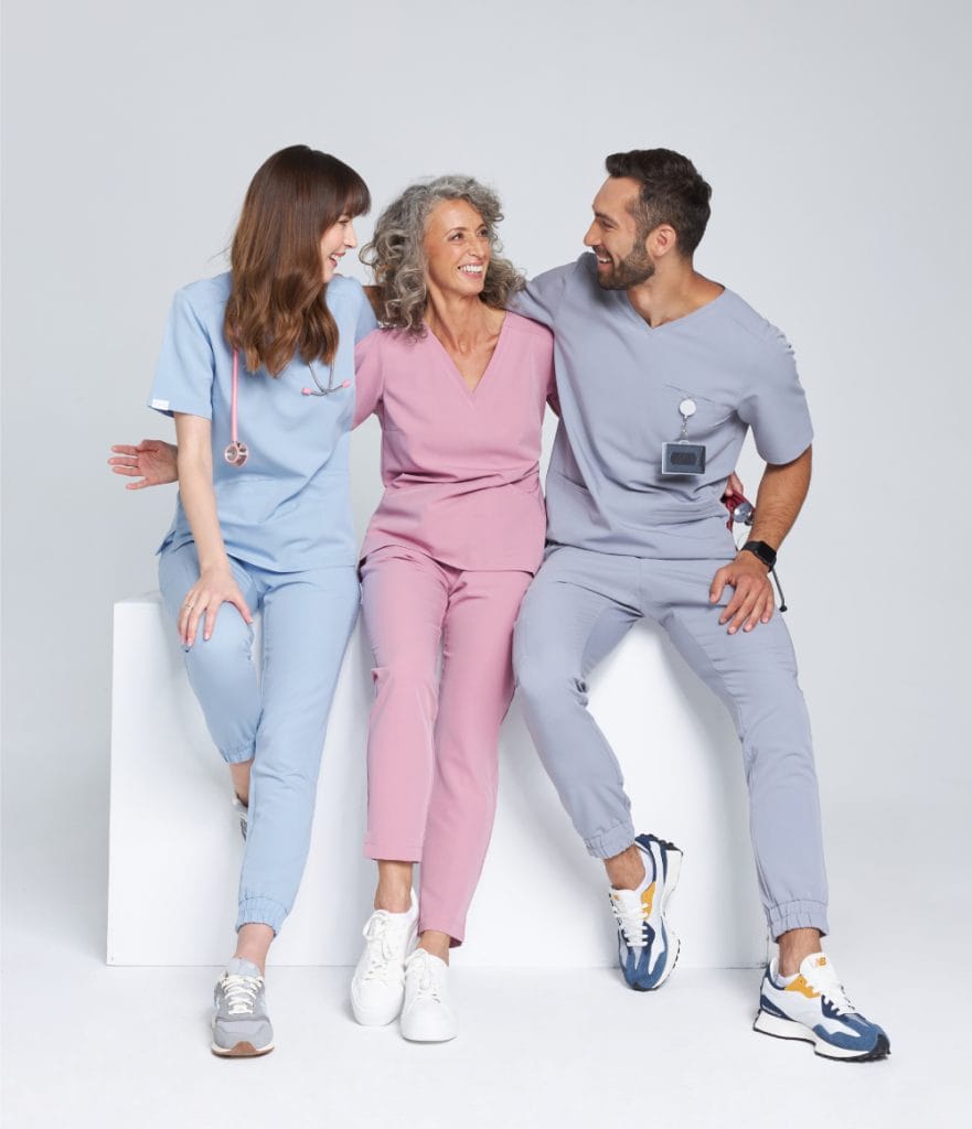 Trzy uśmiechnięte osoby w kolorowej odzieży medycznej siedzące razem na białym bloku.