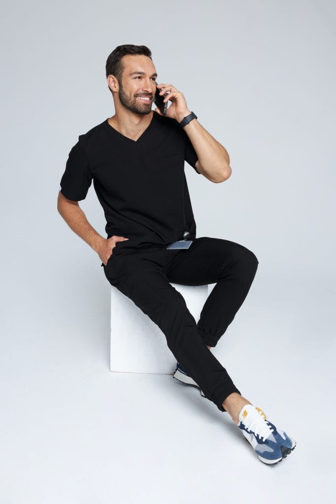 Mężczyzna w czarnej męskiej bluzce medycznej scrubs Basic rozmawia przez telefon.