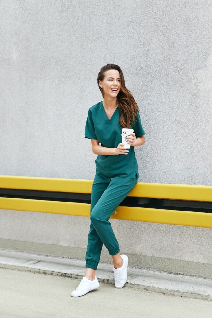 Kobieta w butelkowozielonych spodniach medycznych joggerach i bluzie medycznej trzyma kubek z kawą