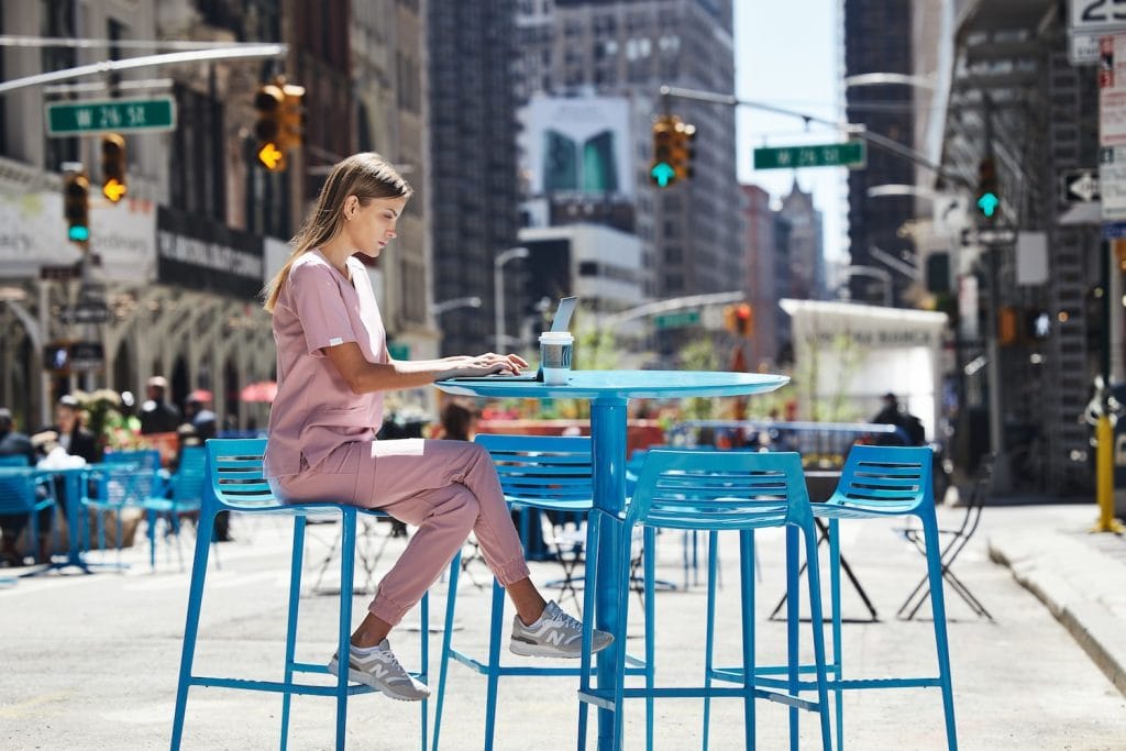 Kobieta w różowym ubraniu medycznym pracuje na laptopie przy niebieskim stole na zewnątrz w miejskim otoczeniu.