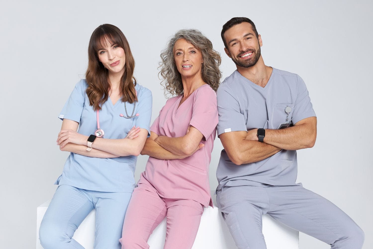 Trzy uśmiechnięte osoby w kolorowej odzieży medycznej pozujące na białym tle.