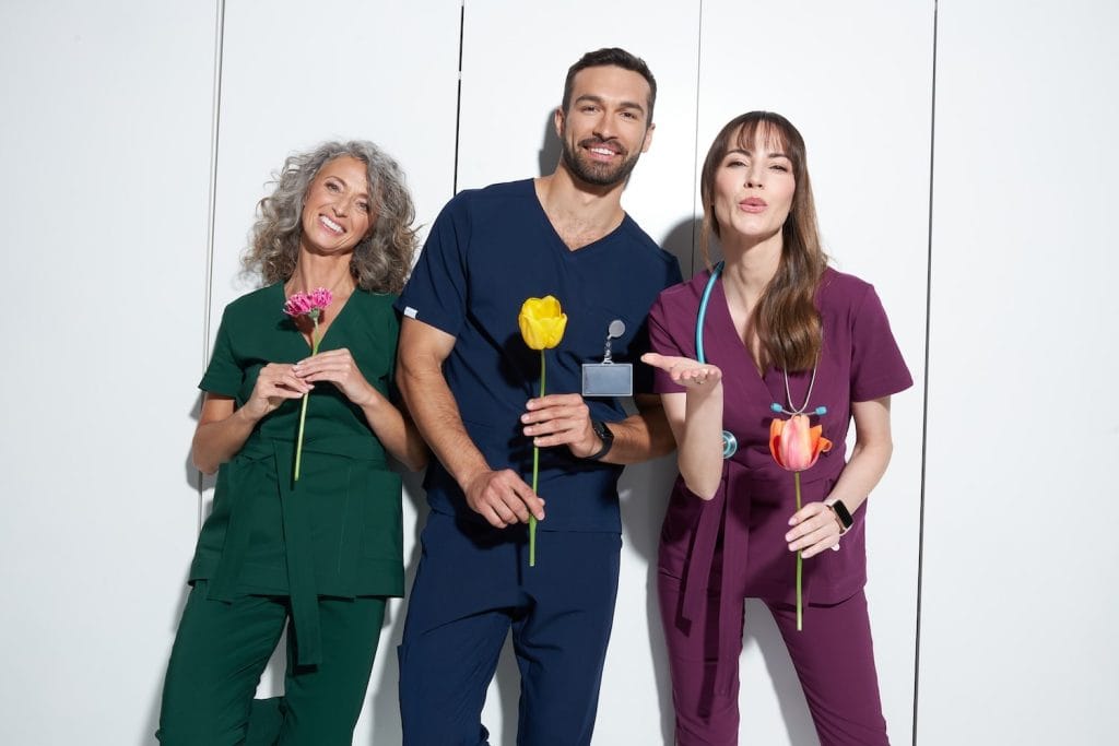 Trzy uśmiechnięte osoby w wielokolorowej odzieży medycznej trzymające kwiaty.