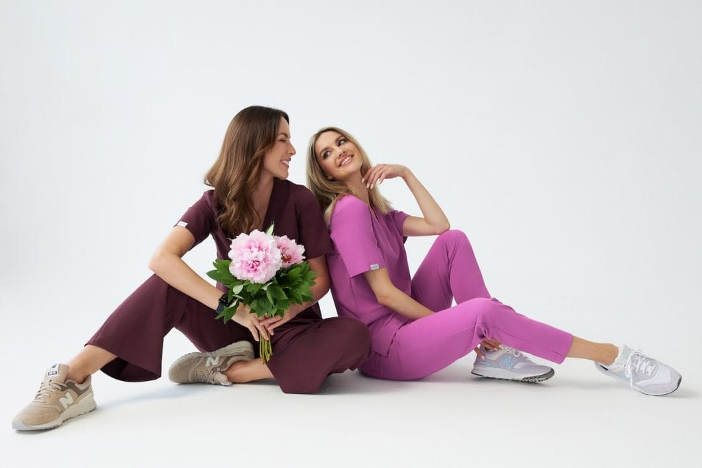 Dwie kobiety w kolorowej odzieży medycznej siedzące na podłodze, uśmiechnięte i trzymające kwiaty.