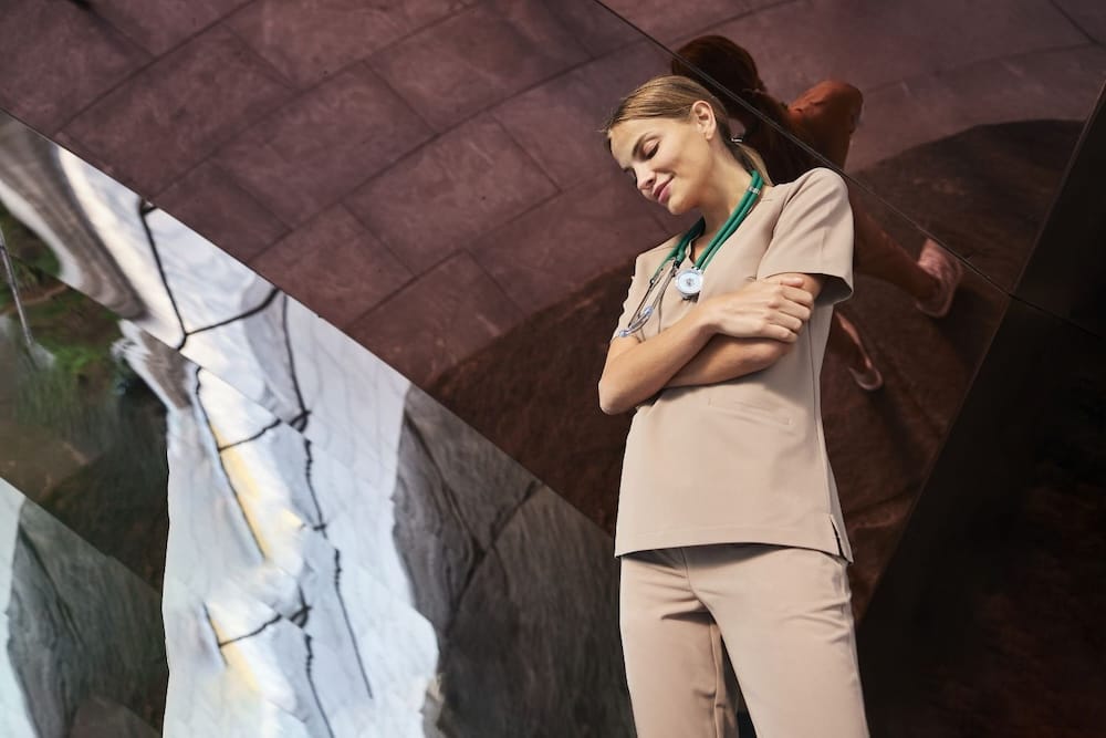 Młoda kobieta w beżowej odzieży medycznej z zielonym stetoskopem stoi z założonymi rękami.